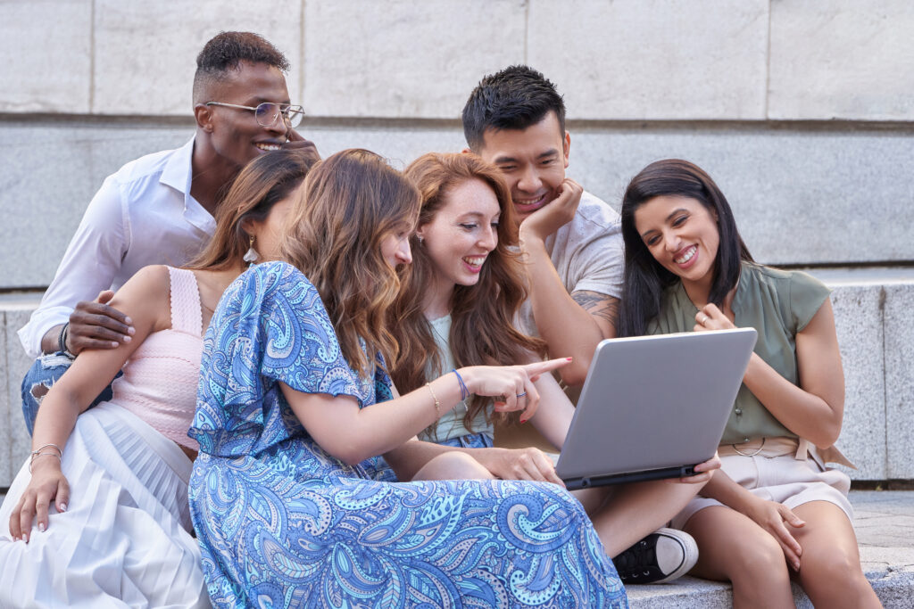 Group of multi-ethnic people using laptop together while sitting on stairs outdoors. Urban lifestyle, technology and team concept.