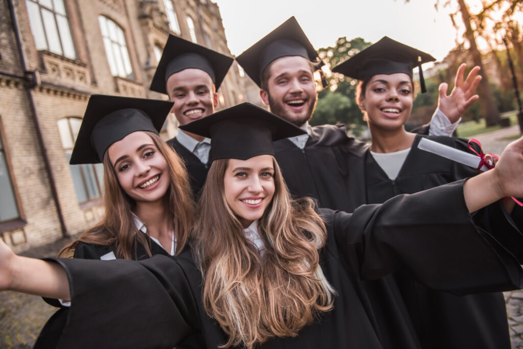 Successful graduates in academic dresses are holding diplomas, looking at camera and smiling while standing outdoors